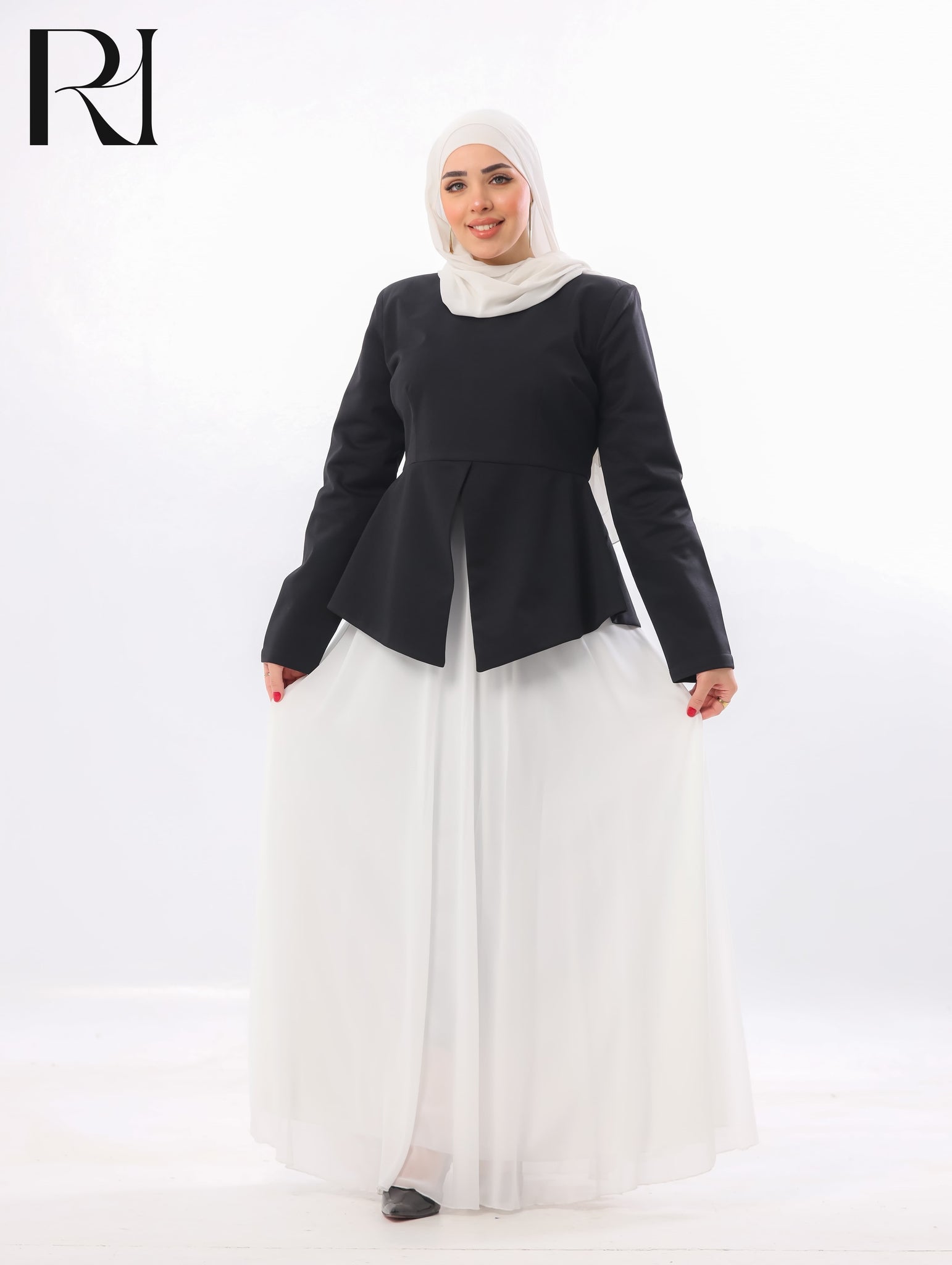 A woman wearing a black long-sleeve peplum-style top and a flowing white maxi skirt, paired with a white hijab, standing against a plain white studio background. A black 'RJ' logo appears in the top-left corner.