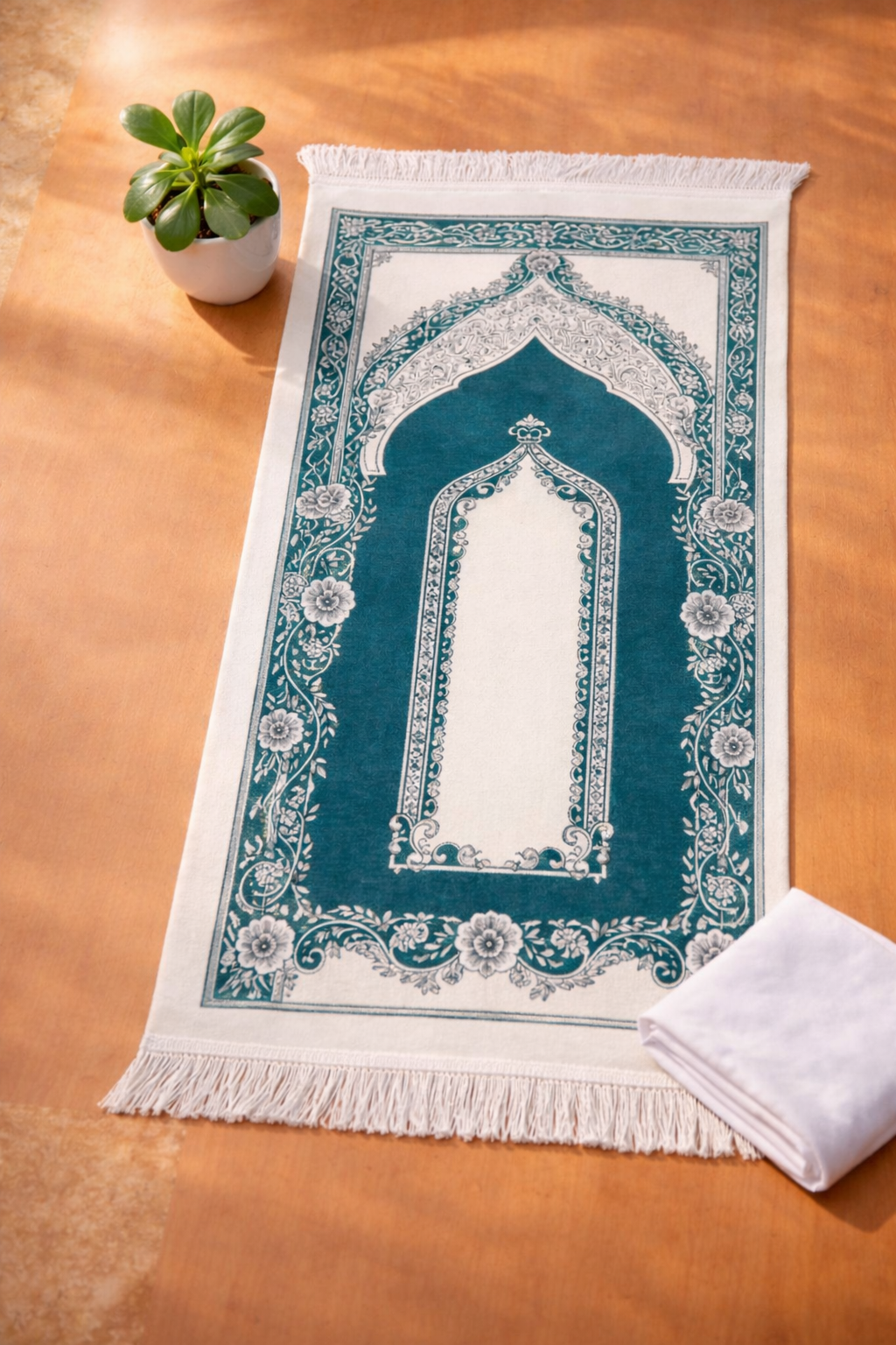 Decorative prayer mat with intricate design on a wooden surface, accompanied by a white towel and small potted plant.