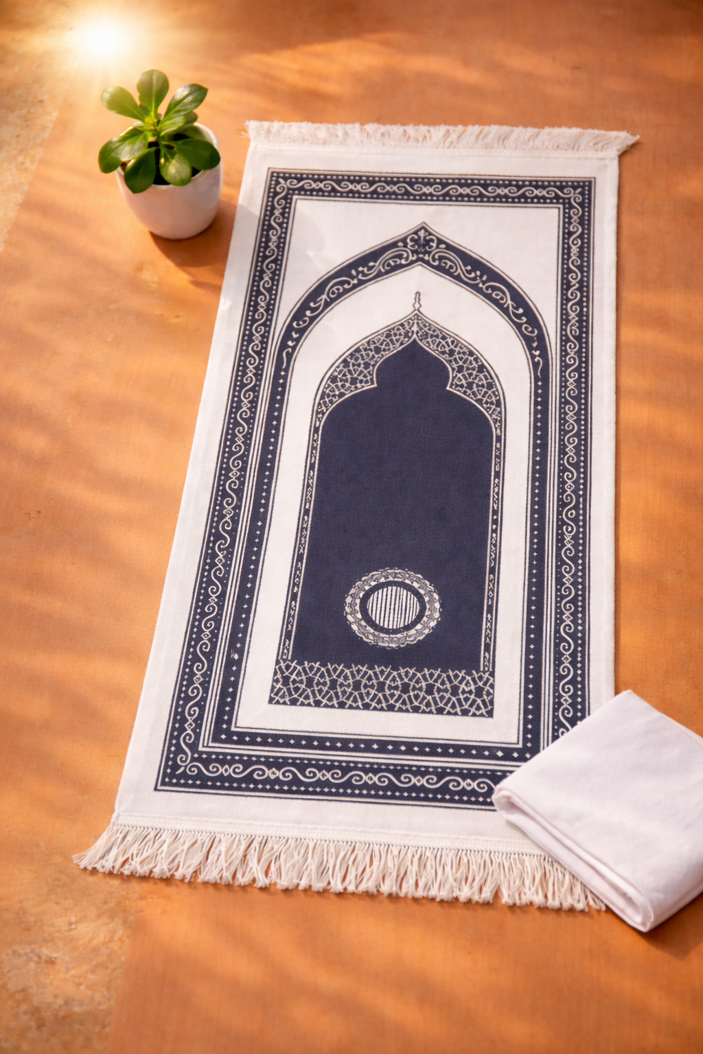 Navy prayer mat with white border on a wooden floor, accompanied by a white towel and small potted plant.